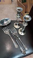 Whole set of silverplated ladle, two ornate serving forks, and pair of candle holders displayed on black surface, showing overall condition and design.