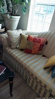 Striped pale lemon yellow and blue sofa with various throw pillows including beige, yellow, multicolor patchwork, and blue pillows, positioned near window and plant.