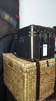Front view of the antique style black box suitcase with metal corner reinforcements sitting on the wicker basket.