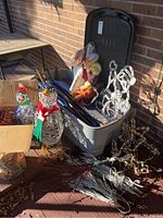 Photo showing nativity figures inside partially open grey plastic storage bin along with lighted wire penguin sculptures and various yard stakes.