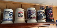 Front view of 5 Denver Broncos beer steins, including white ceramic, black ceramic, and two tall glass steins.