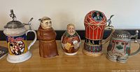 Five figural beer steins arranged in a row against a white wall, showing different designs such as Planters graphic, monks, hot air balloon, and lederhosen.