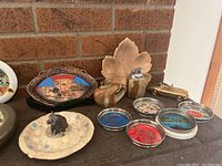 Photo showing various vintage lighters, ashtrays with decorative ceramic and metal elements, and a brass model car on a dark wooden surface against a brick wall.