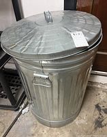 Front angle of a metal trash bin with a round handle lid and side latch handles, showing wear and dirt on the surface.