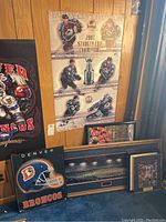 Photo shows several sports posters including 2001 Stanley Cup Champions, Denver Broncos helmet poster, framed stadium print, and small framed sports photos.