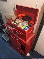Red metal tool chest cabinet in basement, door off, top lid open showing contents inside including hand tool and various hardware.