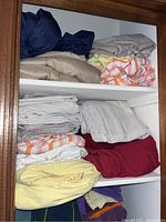 Closet shelf with folded bed sheets and pillowcases in navy, beige, cream with orange stripes, yellow, red.