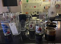 Overview showing approximately 20 vintage brewery and sports-themed pint glasses and mugs on a black surface with multiple detailed logo designs visible.