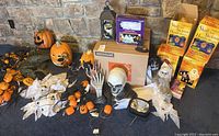 Wide view of vintage Halloween plastic string lights with skull and skeleton hands, blow mold jack-o-lantern pumpkins, ghost figure, and various Halloween decorations on floor against stone wall.