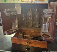 Full view of vintage wooden liquor tantalus set with two doors open showing glass decanters and shot glasses, drawer open showing bar tools.