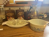 Photo showing vintage Pyrex blue floral mixing bowl, glass canisters labeled Sugar and Flour with wooden lids, glass coffee server with metal stand and punch bowl with cups in background.