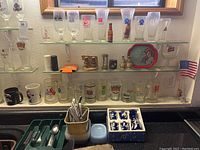 Overview of multiple vintage beer steins, various branded pint glasses, flatware in utensil holders, small sake set dishes, and collectibles placed on shelves and kitchen counter.