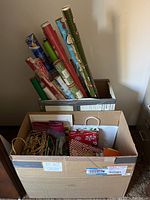 Two large cardboard boxes filled with Christmas wrapping paper rolls and various gift bags with greeting cards and envelopes.