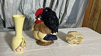 Photo showing ceramic rooster, yellow decorative vase with floral motif, and round seascape wall art on a white cloth surface