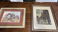 Two framed prints on a wooden table showing farmhouse and cityscape scenes.