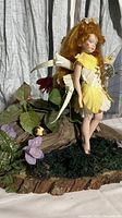 Garden nymph doll with red hair and yellow and white petal dress standing barefoot on a natural wood slice base covered with green moss and surrounding faux flowers and plants