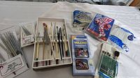 Photo of assortment of artist brushes in boxes alongside painting accessories on a white cloth.
