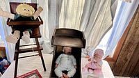 Cabbage Patch doll seated in wooden high chair next to another wooden high chair with vinyl baby doll lying in it and another baby doll sitting. Curtain background with natural lighting.
