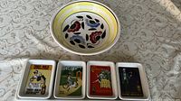 Photo of a round hand-painted ceramic bowl with floral and abstract patterns placed behind four rectangular serving dishes with vintage graphic advertisements.