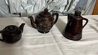 Three pottery pieces on white cloth: two teapots and a pitcher, all dark brown glazed pottery. One teapot with floral painted decoration visible.