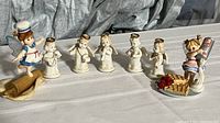 Wide view showing all seven white Japanese angel figurines in a row and two Little Kitchen Fairies figurines at either end on a white cloth background