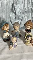 Five vintage bisque figurines arranged on a white cloth backdrop showing front views and details