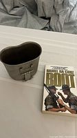 Vintage marine canteen cup made of metal with typical shape, placed next to the paperback book on a white surface.