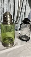 Two vintage look candle lanterns, a black Yankee Candle lantern on the right and a green glass lantern with metal top on the left, displayed upright on a white fabric background.