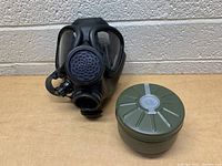 Front view of black military-style gas mask placed next to green filter canister on a wooden surface with a cinder block wall background.