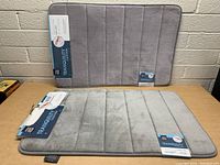 Two memory foam bath mats laid flat on a wooden surface against a wall, both tagged as new from the Tranquility Collection