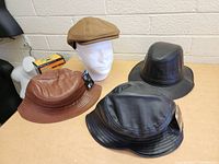 Lot of 4 hats shown together on a table, including two black leather flat caps, one brown leather bucket hat, and one tan fabric flat cap on a white mannequin head.
