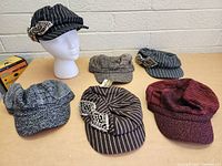 Front view of six women's hats on display table, showing different patterns and brooch decorations on two black pinstripe caps and a red beret