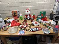 Full view of assorted Christmas items including figurines, stockings, ornaments, mugs, plates, and decorations on a table against a white brick wall.