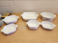 All six CorningWare dishes with blue cornflower design and one plastic lid arranged on table.