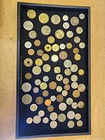 Top-down photo of approximately 60 foreign coins arranged in a black felt tray showing a variety of sizes, metals, and some with holes in the center.