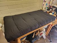 Black cushioned seat pad lying flat on wooden table. Visible tufting and stitching detail. New condition with no stains or wear visible.