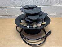 Front view of the small indoor water fountain showing the layered slate stones, decorative polished stones, base with Greek key pattern, and power cord