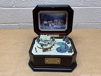 Front angle showing the open music box with winter scene including snow-covered house, horse-drawn sleigh, ice skating pond, and pine trees inside wooden case