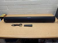 Front view of Wohome Bluetooth soundbar with remote and AUX cable on a brown table against a brick wall.