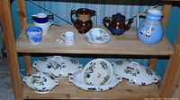 Overview of multiple china items on wooden shelves showing jugs, cups, teapots, and Victorian tureens and dishes with butterfly and botanical motifs