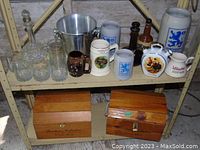 Overview photo showing assortment of barware items including German stoneware steins, glass decanters, etched ship glass tumblers, metal ice bucket, brown ceramic container, and two wooden wine boxes.