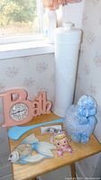 Photo showing pink bath clock spelling 'Bath' with clock face, blue and white knitted poodle toilet paper holder, tall white plastic toilet paper holder, pastel chalk fish, and girl wall pocket with repaired bow on small wooden table in bathroom setting.