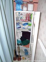 View of a white bathroom cabinet containing various toiletries and linens, including shampoo bottles, hand sanitizer, cotton balls, towels, face cloths, and plastic containers with white powders.