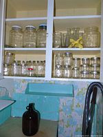 Photo of cabinet shelves filled with multiple clear glass canning jars with metal lids and a brown glass jug below on teal tiled counter.