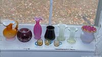Seven collectible glass pieces displayed on a windowsill, showing variety in colors and shapes, including vases and bowls.