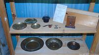 Wide view of wooden shelves holding assorted brass and copper dishes, small cast iron pot, a wooden carved netsuke, a Scottish spoon, and a brown vintage bank coin holder shaped like a book
