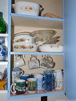 Blue cupboard shelf showing Corningware casserole dish with floral decoration, glass lids, ceramic and glass mugs in varied designs and colors.