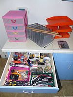 Drawer open showing assorted office supplies like scissors, pens, rulers, clips, small baskets with tacks, staples, and various items alongside multiple organizers including pink drawer organizer, metal mesh file sorter, orange paper trays, and calculator on countertop.