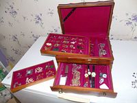 Open jewelry chest showing multiple trays with various jewelry pieces including earrings, brooches, watches, and pins on red fabric lining