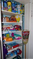 Shelf of assorted cleaning liquids, sprays, textiles including dish cloths, towels, aprons, rags, a corn broom leaning against shelf.